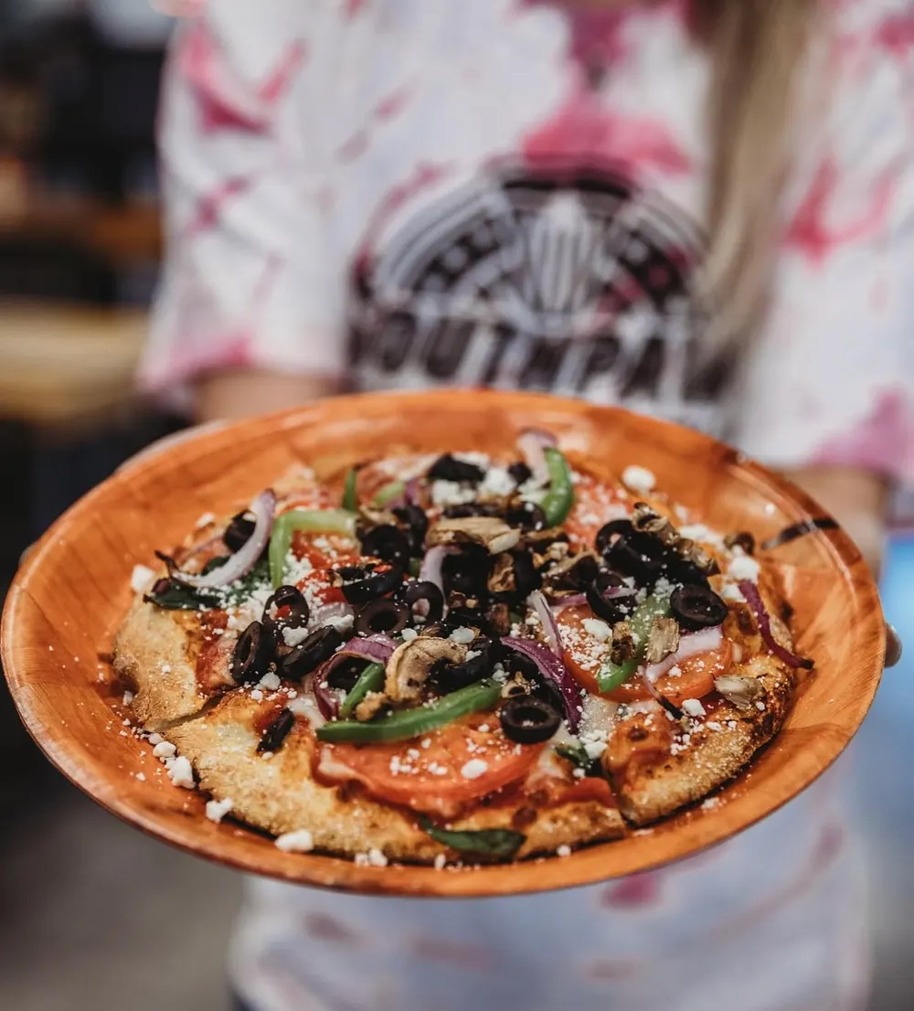A pizza held out on a wooden serving tray by someone in a Southpaw's shirt